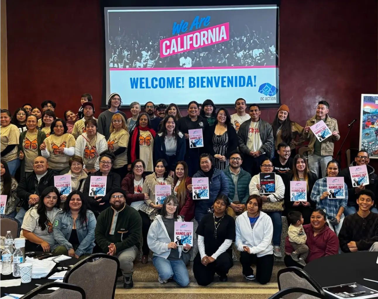 A crowd of people holding signs, arranged in tiers in front of a projector screen that says "We Are California."